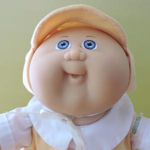 May include: A doll with a round face, bright blue eyes, and a small nose. The doll wears a pale yellow cap, a white collared shirt with a tie-front closure, and a yellow garment. A small green tag is visible on the sleeve of the white shirt.
