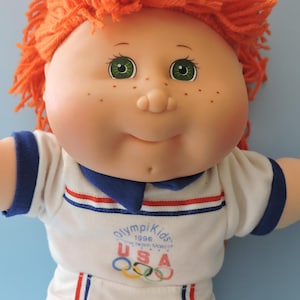 May include: A vintage Cabbage Patch Kids doll with orange yarn hair, green eyes, and freckles. The doll wears a white shirt with blue and red stripes and the text "Olympikids 1996 USA" with Olympic rings.