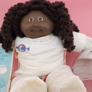 May include: A Cabbage Patch Kids Young Astronaut doll with brown yarn hair, wearing a white astronaut suit with a patch that says "Astronauts". The doll has a birth certificate and adoption papers.