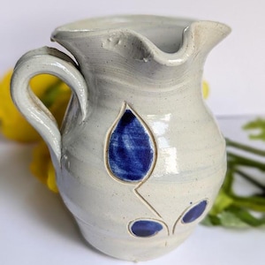 May include: A small white ceramic pitcher with a blue and white floral design. The pitcher has a handle and a spout.
