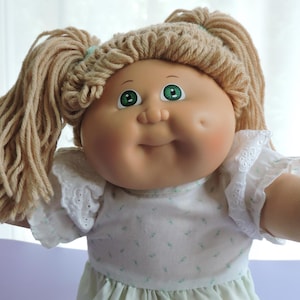 May include: A doll with light tan skin, green eyes, and dimpled cheeks. The doll has blonde yarn hair styled in two high pigtails with light blue hair ties. It wears a white short-sleeved dress with eyelet lace trim on the sleeves and a small pattern.