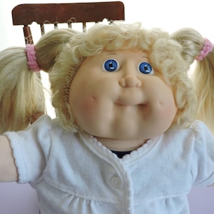 May include: A vintage Cabbage Patch Kid doll with blonde hair styled in pigtails secured with pink scrunchies. The doll has blue eyes, rosy cheeks, and a smiling expression. It is wearing a white, button-up sweater.