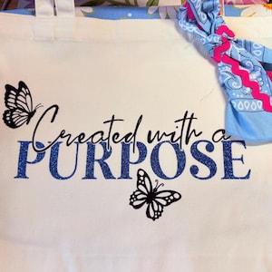 Puede incluir: Bolso de mano blanco con el texto "Created with a PURPOSE" en escritura negra y letras brillantes azules. Dos ilustraciones de mariposas negras están en la bolsa. Un pañuelo azul con ribete rosa está atado al asa.