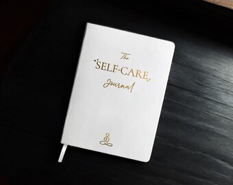 May include: White journal with gold lettering that reads "The Self-Care Journal" and a gold symbol of a person sitting in a meditative pose.