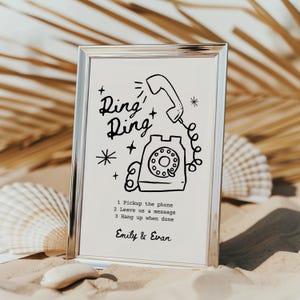 May include: A silver frame with a white background and a black and white illustration of a rotary phone with the text "Ring Ring" and instructions to "Pickup the phone, Leave us a message, Hang up when done". The names "Emily & Evan" are printed at the bottom of the frame.