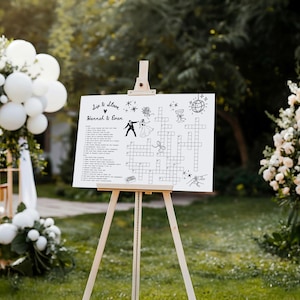 May include: A white sign on a wooden easel with the words "Sip & Solve" and "Hannah & Evan" in black script. The sign features a crossword puzzle with wedding-themed clues and illustrations. The easel is made of light-colored wood and is set outdoors.