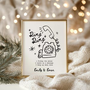 May include: A black and white illustration of a rotary phone with the text "Ring+Ring" above it. The illustration is on a white background with a gold frame. The text below the illustration reads "1 Pickup the phone 2 Leave us a message 3 Hang up when done" and "Emily & Evan".