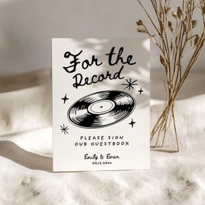 May include: A white sign with black text and a black and white illustration of a vinyl record. The text reads "For the Record" with stars and the words "Please sign our guestbook" below the record. The names "Emily & Evan" and the date "09.12.20XX" are at the bottom of the sign.
