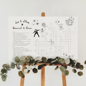 May include: A white wedding crossword puzzle sign with the names "Hannah & Evan" in script. The sign includes a crossword puzzle with clues, illustrations of a bride and groom, and decorative elements.
