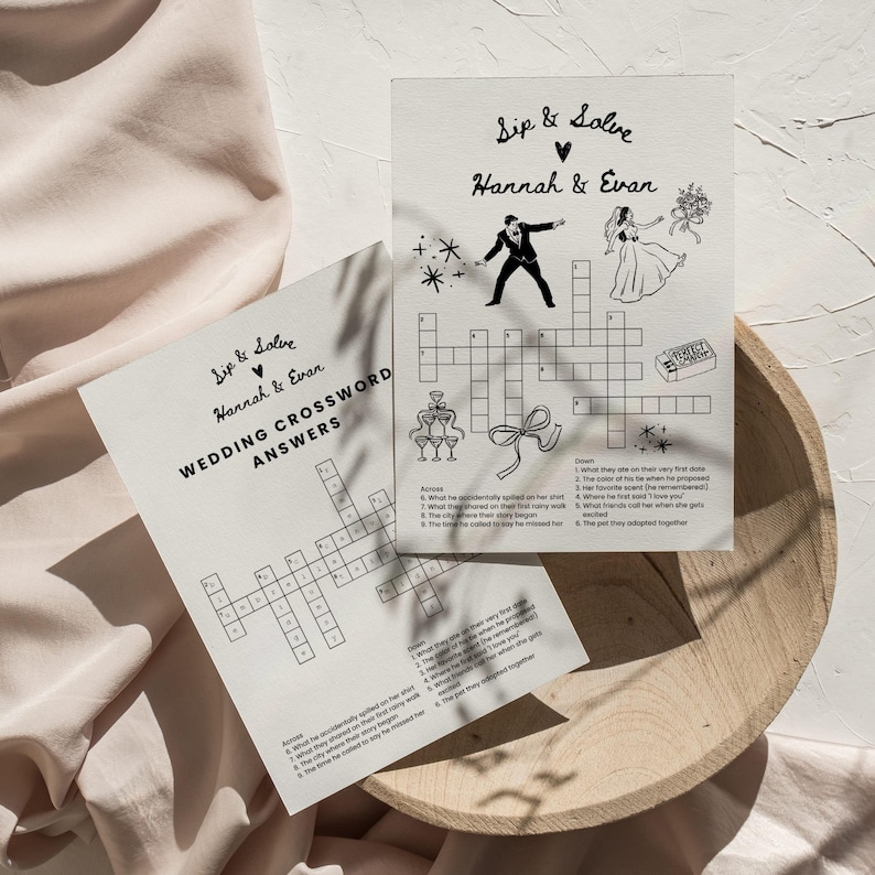 May include: Two wedding-themed crossword puzzles on a wooden tray. One puzzle is labeled "Wedding Crossword Answers" and the other is a crossword puzzle with the text "Sip & Solve Hannah & Evan". The puzzles are printed in black ink on off-white paper.