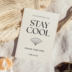 K&ouml;nnte beinhalten: Eine cremefarbene Karte mit den Worten "STAY COOL" in gro&szlig;en schwarzen Buchstaben. Unter dem Text befindet sich eine F&auml;cherillustration. Die Karte enth&auml;lt auch die S&auml;tze "TO OUR BIGGEST FAN" und "PLEASE TAKE ONE!" mit den Namen "Emily & Evan" unten.