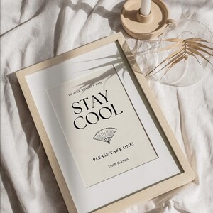 K&ouml;nnte beinhalten: Ein gerahmter Druck mit den schwarzen Worten "STAY COOL" und einer F&auml;cherillustration. Der Text "TO OUR BIGGEST FAN" und "PLEASE TAKE ONE!" sind ebenfalls enthalten. Der Rahmen ist aus hellem Holz und der Hintergrund ist neutral.