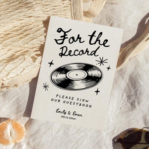 May include: A white sign with black text and a black and white illustration of a vinyl record. The text reads "For the Record" and "Please sign our guestbook." The sign also includes the names "Emily & Evan" and the date "09.12.20XX".