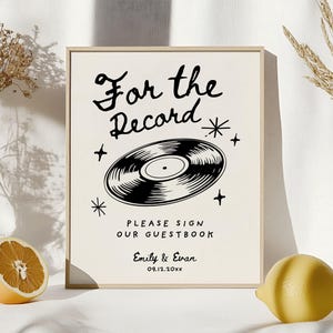 May include: A white sign with a black vinyl record illustration and the text "For the Record". Below the record is the text "Please sign our guestbook" and the names "Emily & Evan" with the date "09.12.20xx".