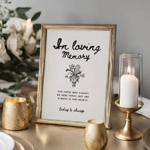 May include: A framed sign with the words "In loving Memory" in black script, along with a floral illustration. The sign is in a gold frame, and there is a lit candle in a glass holder to the right.