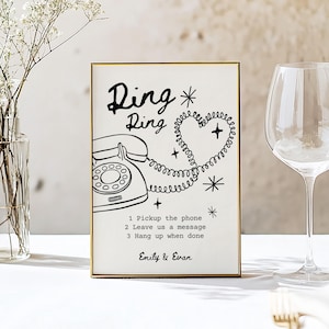 May include: A framed sign with a gold border features a black and white illustration of a vintage telephone and the words "Ring Ring". The text includes instructions and the names "Emily & Evan". The design incorporates a heart-shaped phone cord.