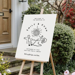 May include: A cream-colored wedding welcome sign on a wooden easel. The sign features black text and a line drawing of a couple dancing under a disco ball. Text includes the names "Emily & Evan", the date, location, and the phrase "We're glad you are here."