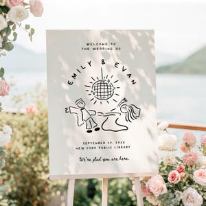 May include: A white wedding welcome sign on an easel. The sign reads "Welcome to the Wedding of Emily & Evan" with a disco ball illustration. The date and location are also listed. The sign is surrounded by flowers.