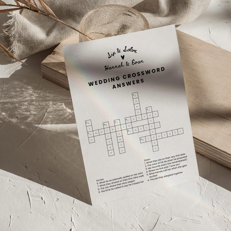 May include: A wedding crossword puzzle with the text "Sip & Solve, Hannah & Evan, Wedding Crossword Answers." The crossword has clues and answers related to the couple's story. The paper is on a wooden surface.