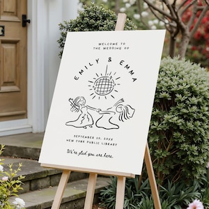 May include: A cream-colored wedding welcome sign on a wooden easel. The sign features black text and illustrations, including the names "Emily & Emma", a disco ball, and two dancing figures. The date and location are also displayed.