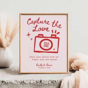 May include: A pink and white sign with a gold frame. The sign says "Capture the Love" in red text with a red cartoon camera and a QR code. The text below the camera says "Share your photos with us! Simply scan and upload." The sign is also signed "Emily & Evan" and the date "November 11, 2025".