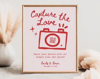 May include: A pink and white sign with a gold frame. The sign says "Capture the Love" in red text with a red cartoon camera and a QR code. The text below the camera says "Share your photos with us! Simply scan and upload." The sign is also signed "Emily & Evan" and the date "November 11, 2025".