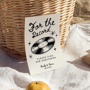 May include: A white sign with black text that reads "For the Record" with a black and white illustration of a vinyl record. The text below the record says "Please sign our guestbook" followed by "Emily & Evan" and the date "09.12.20xx".