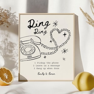 May include: A framed print with a cream background features a black illustration of a vintage rotary phone. The text "Ding Ring" is above the phone, with a heart-shaped phone cord. Instructions and names are below.