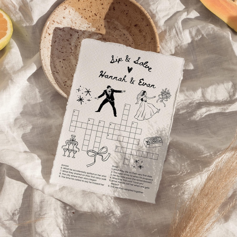 May include: A handmade paper wedding crossword puzzle with the text "Sip & Solve Hannah & Evan". The black and white illustration includes a bride, groom, champagne glasses, and a bow. The puzzle is on a white surface.