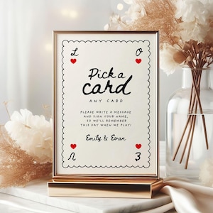 May include: A framed sign with the text "Pick a card" in black script. The sign has a gold frame and a white background with red hearts and the names "Emily & Evan". The sign is on a gold stand.