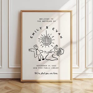 Puede incluir: Un letrero de boda enmarcado con un marco dorado. La ilustración en blanco y negro presenta una bola de discoteca y figuras bailando. El texto incluye los nombres "Emily & Evan", la fecha, el lugar y la frase "We're glad you are here."