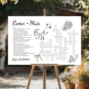 May include: A white wedding crossword puzzle on an easel, featuring the names "Evan + Mia" at the top. The puzzle includes clues and spaces for answers, with decorative illustrations of a couple, flowers, and champagne glasses.