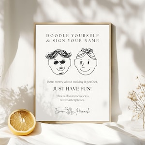 May include: A framed print with the text "DOODLE YOURSELF & SIGN YOUR NAME" and "JUST HAVE FUN!". The print features two cartoon faces and is signed "Evan & Hannah". A halved lemon sits in front of the print.