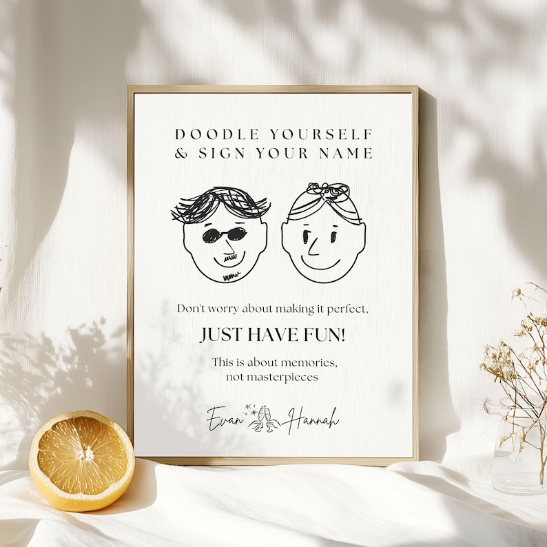 May include: A framed print with the text "DOODLE YOURSELF & SIGN YOUR NAME" and "JUST HAVE FUN!". The print features two cartoon faces and is signed "Evan & Hannah". A halved lemon sits in front of the print.