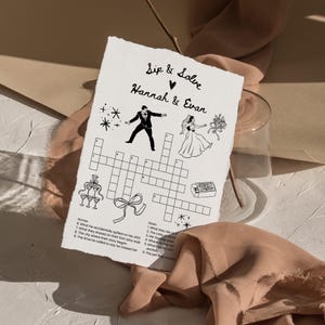 May include: A wedding-themed crossword puzzle on textured paper. The puzzle is titled "Sip & Solve Hannah & Evan" and features illustrations of a bride, groom, champagne glasses, and a bow. The crossword has clues and answers related to the couple's story.