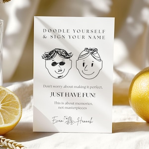 May include: A white card with black text and doodle illustrations. The text reads "DOODLE YOURSELF & SIGN YOUR NAME" and "JUST HAVE FUN!". Below the text are two doodle faces and the names "Evan" and "Hannah".
