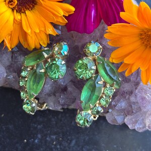 May include: A pair of gold-tone earrings featuring clusters of light green and dark green faceted rhinestones in marquise and round shapes. The earrings are arranged in a curved, leaf-like design with prong settings holding each individual stone.