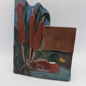 May include: A decorative, rectangular wall plaque featuring a carved duck scene. The plaque depicts a mallard duck on a blue pond, cattails, and a brown rectangular space. The color palette includes shades of brown, green, and blue.