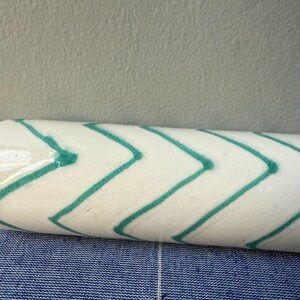 Op de afbeelding: Een witte keramische roller met een turquoise chevron patroon. De roller is cilindrisch en lijkt een decoratief item te zijn. De achtergrond is een blauw-wit patroon.