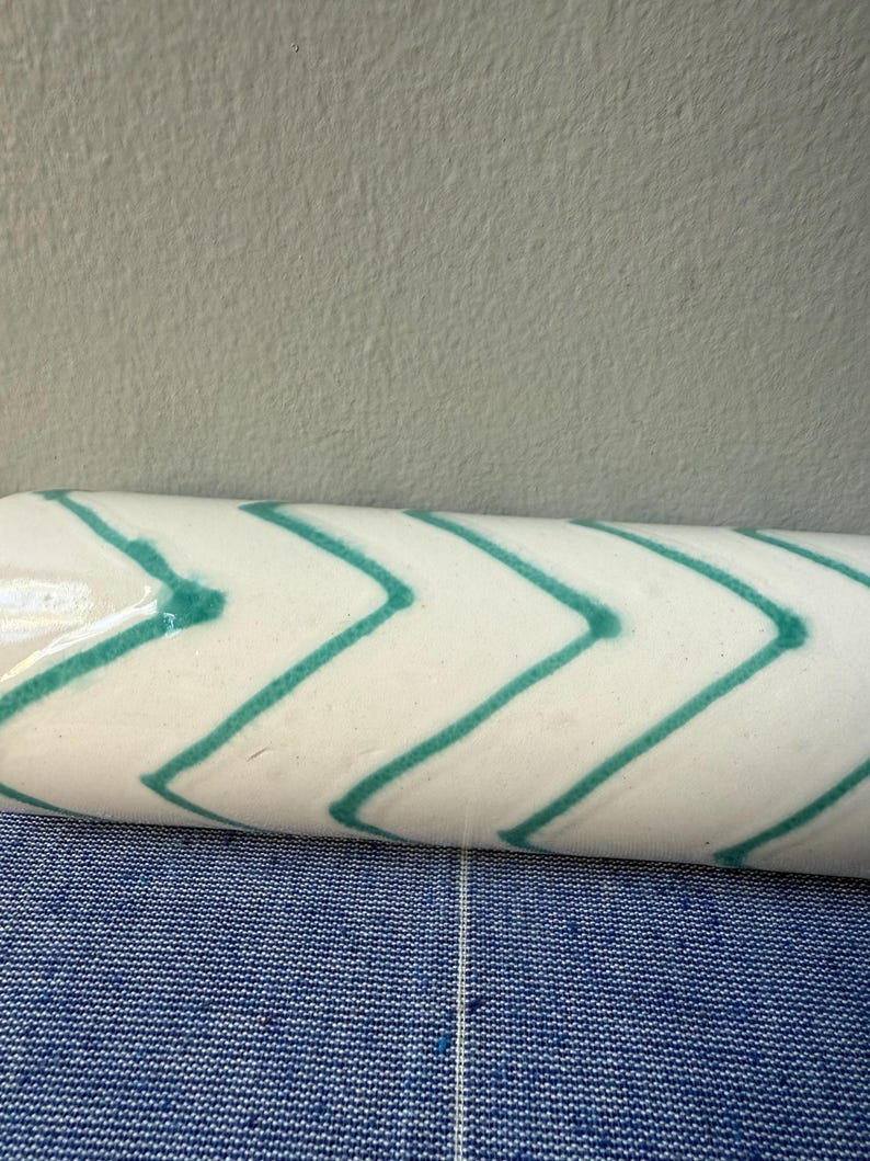 Op de afbeelding: Een witte keramische roller met een turquoise chevron patroon. De roller is cilindrisch en lijkt een decoratief item te zijn. De achtergrond is een blauw-wit patroon.