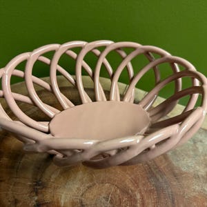 May include: A light pink ceramic bowl featuring a woven, lattice-style openwork design. The bowl has a smooth, glossy finish and a solid, circular base, with curved, interconnected ceramic strips forming the sides of the vessel.