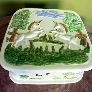 May include: A square ceramic box with a cream-colored base featuring a raised relief design. The lid depicts two white unicorns facing each other in a green landscape with trees and a blue cloud. The sides also feature painted landscape details.