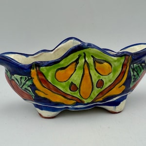 May include: A hand-painted ceramic planter with a vibrant floral design. The planter features a scalloped rim and three small feet. The exterior is decorated with a mix of colors, including blue, green, orange, and red.