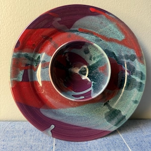 May include: A handmade ceramic serving dish with a central bowl. The dish features a vibrant color palette of red, purple, and teal, with a glossy finish. The design is abstract and artistic, suitable for serving snacks or dips.