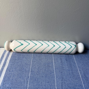 Op de afbeelding: Een witte keramische deegroller met een turquoise zigzagpatroon. De deegroller heeft afgeronde handvatten aan elk uiteinde. Hij ligt op een blauw-wit gestreept oppervlak.