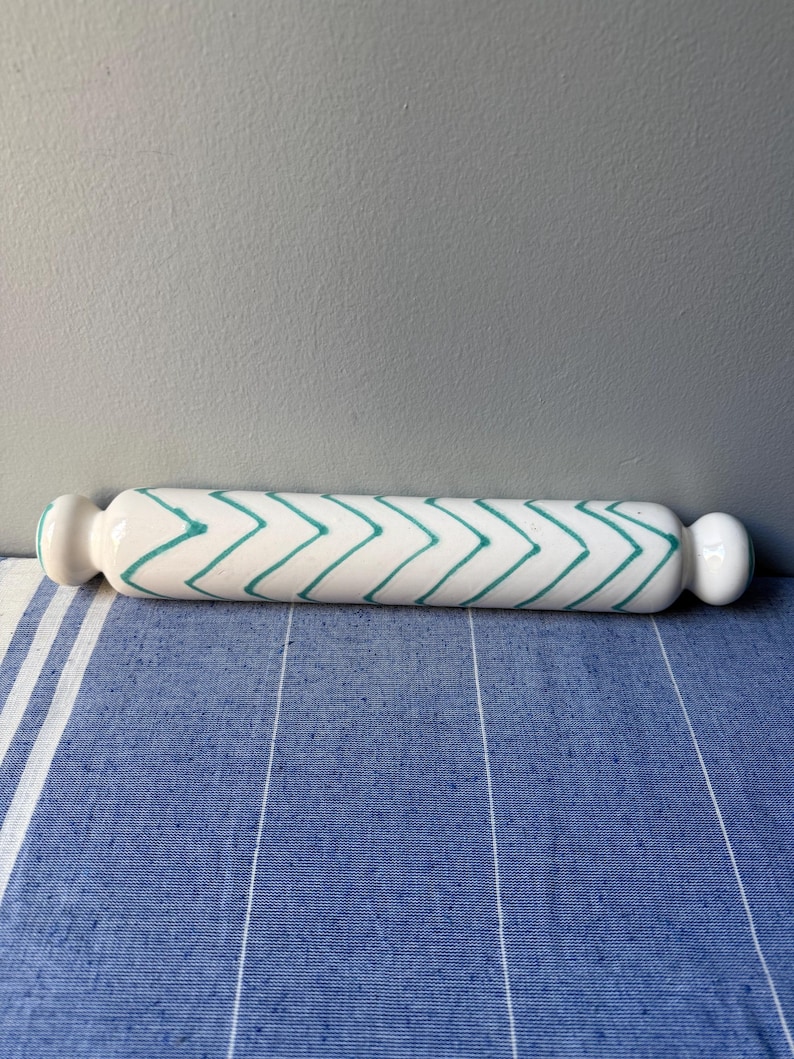 Op de afbeelding: Een witte keramische deegroller met een turquoise zigzagpatroon. De deegroller heeft afgeronde handvatten aan elk uiteinde. Hij ligt op een blauw-wit gestreept oppervlak.