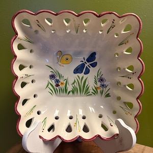 May include: A square ceramic bowl with a scalloped, pink-trimmed edge and lattice-style cutouts. The interior features a hand-painted scene of a yellow butterfly, a blue butterfly, green grass, and small purple, yellow, and blue flowers on white.