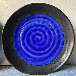 May include: A round ceramic plate with a striking design. The plate features a vibrant blue center with a spiral pattern, framed by a textured black border. The plate's glossy finish enhances the colors, making it a decorative piece for the home.