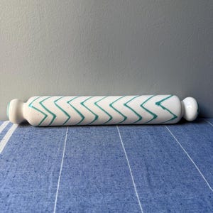 Op de afbeelding: Een witte keramische deegroller met een turquoise zigzagpatroon. De deegroller heeft afgeronde uiteinden en wordt gepresenteerd op een blauw-wit gestreept oppervlak. De achtergrond is effen grijs.