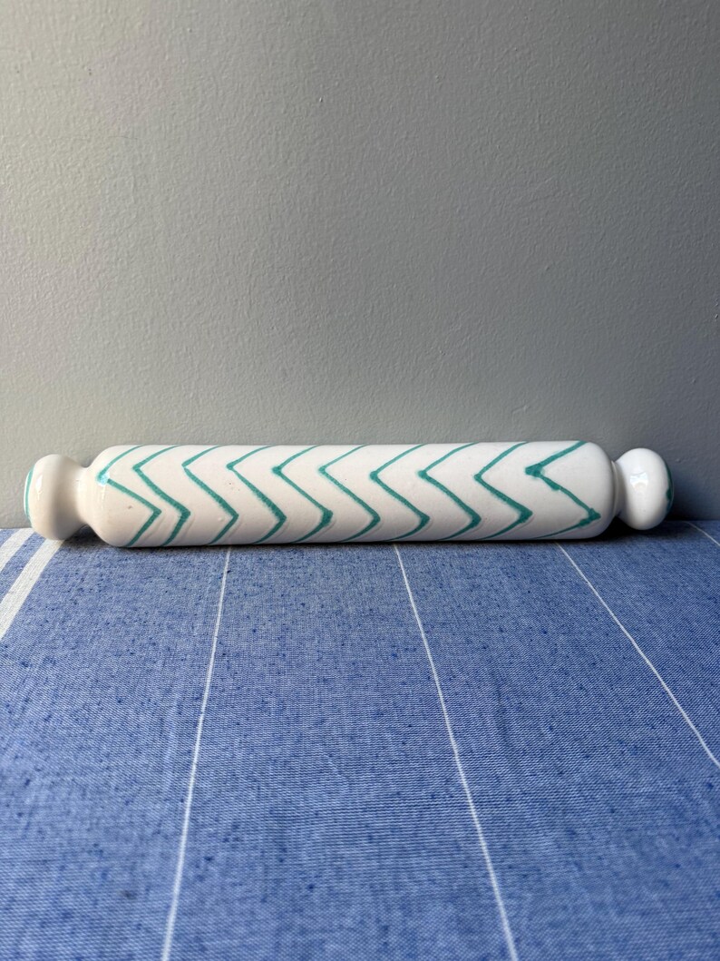 Op de afbeelding: Een witte keramische deegroller met een turquoise zigzagpatroon. De deegroller heeft afgeronde uiteinden en wordt gepresenteerd op een blauw-wit gestreept oppervlak. De achtergrond is effen grijs.